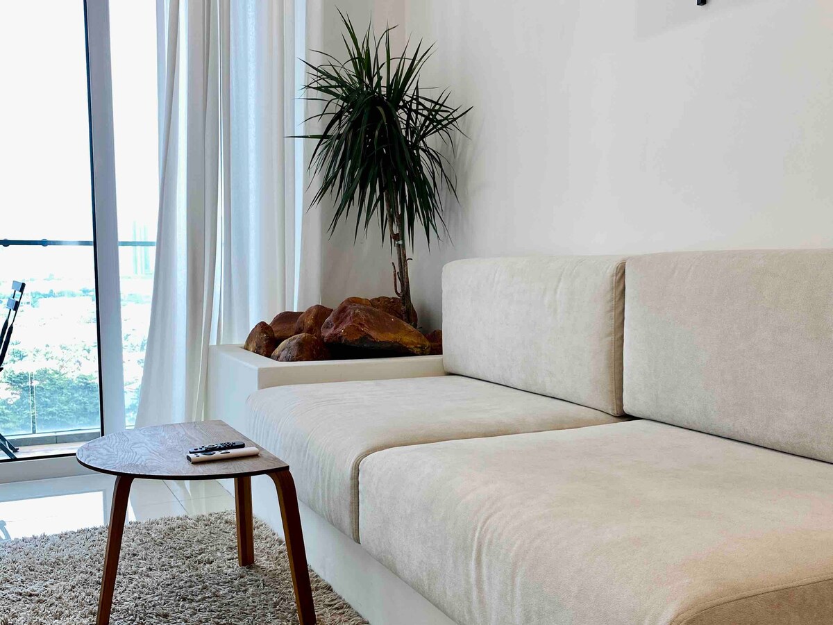 A light, airy living space features a spacious sectional sofa upholstered in a neutral tone, accompanied by a small wooden table. A green potted plant adds natural interest, while soft light filters through sheer curtains, illuminating the area.