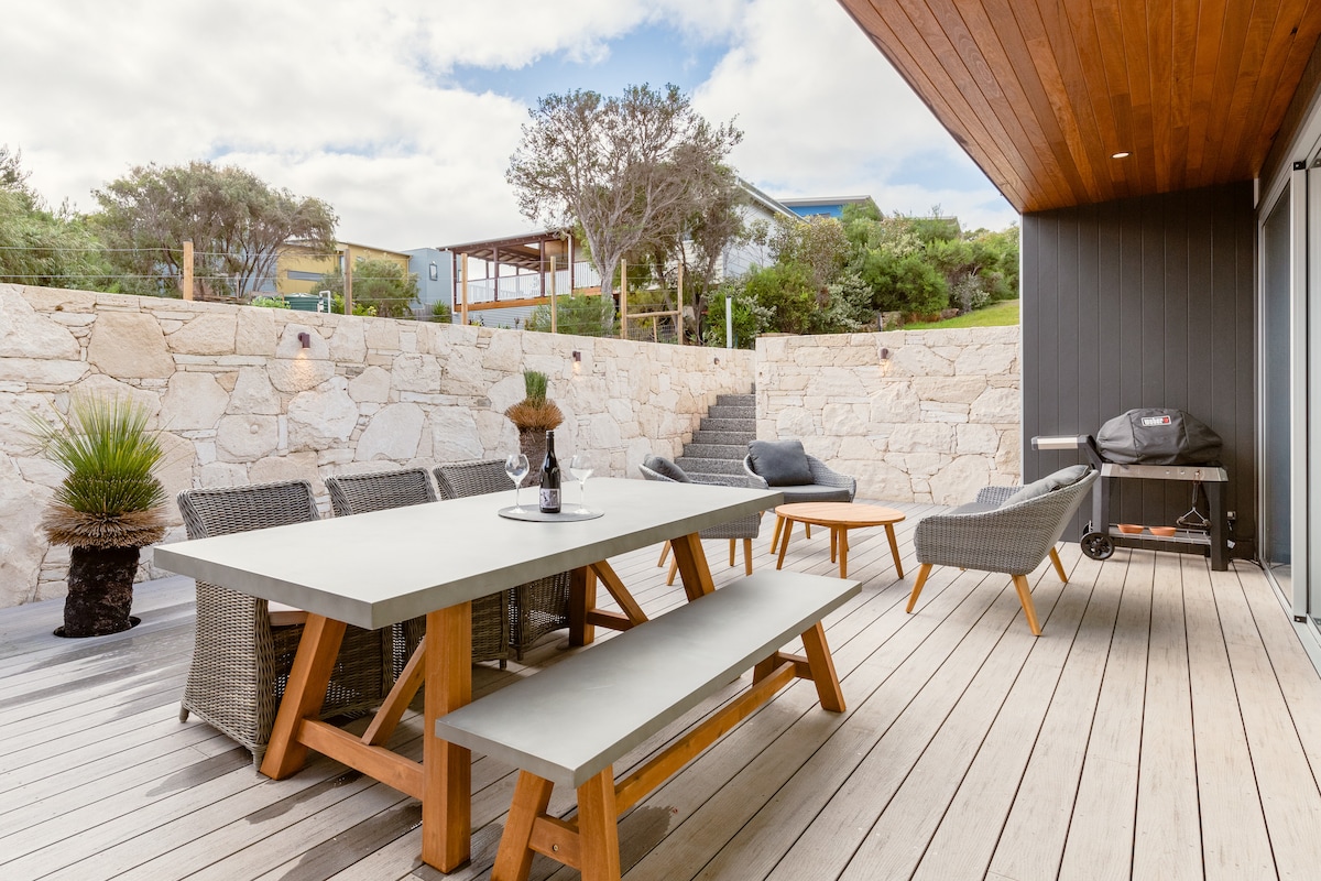 An outdoor dining area is featured with a concrete table surrounded by wooden benches. Lounge chairs are positioned nearby, with a barbecue grill located on a side. Natural stone walls provide a sense of privacy, complemented by lush greenery in the background.
