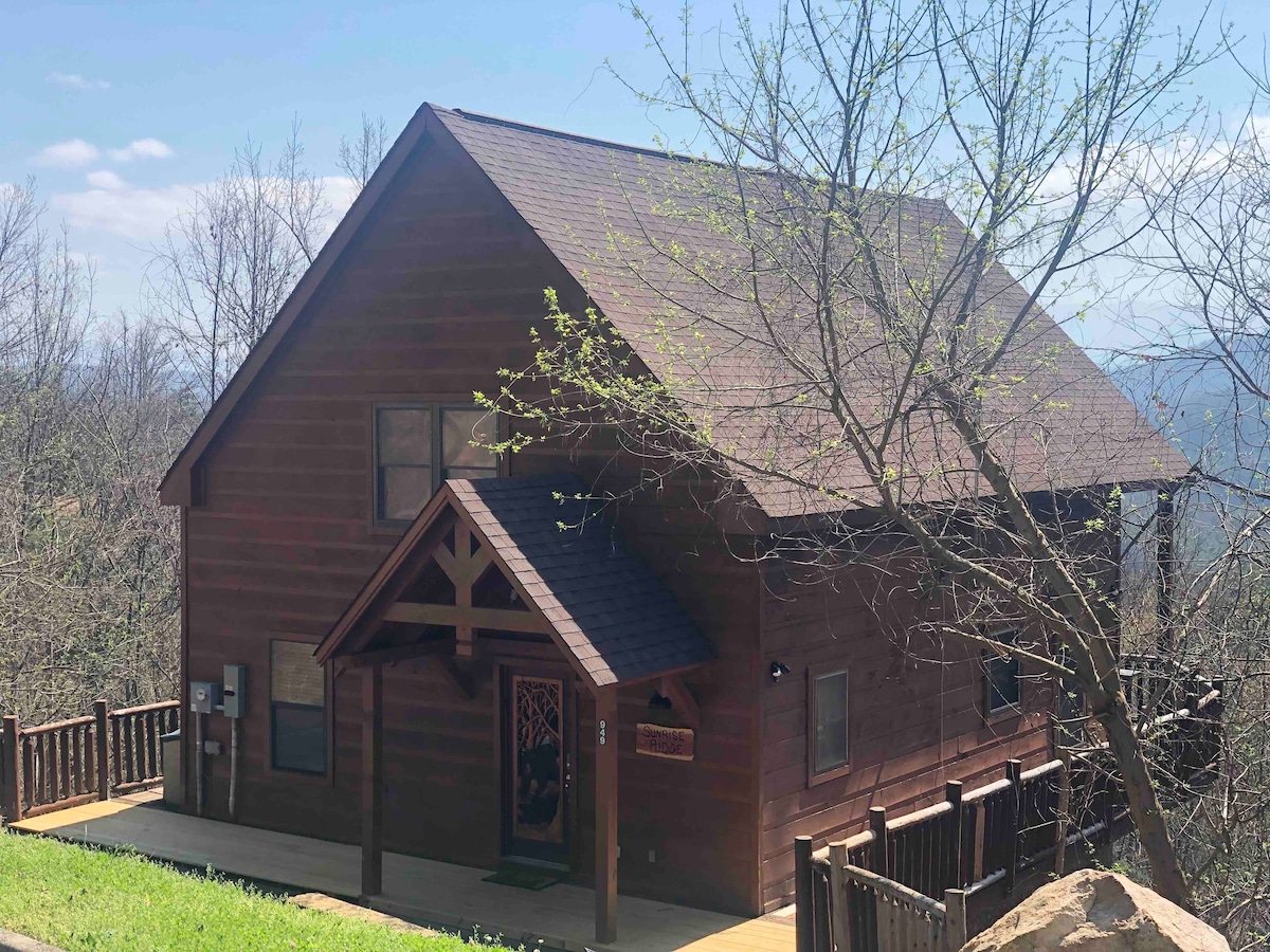 A cozy cabin exterior features rustic wooden siding and a welcoming front porch with a decorative entryway. The two-story structure is surrounded by greenery, enhancing the serene atmosphere. Vast views of the Smoky Mountains can be glimpsed in the background beneath a clear blue sky.