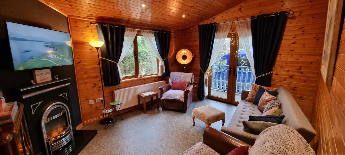 A timber-lined living room features a cozy seating arrangement with a sofa and armchair, paired with a side table. Large windows allow natural light to fill the space, adorned with curtains. A flat-screen TV is mounted on the wall, and a decorative lamp adds warmth.