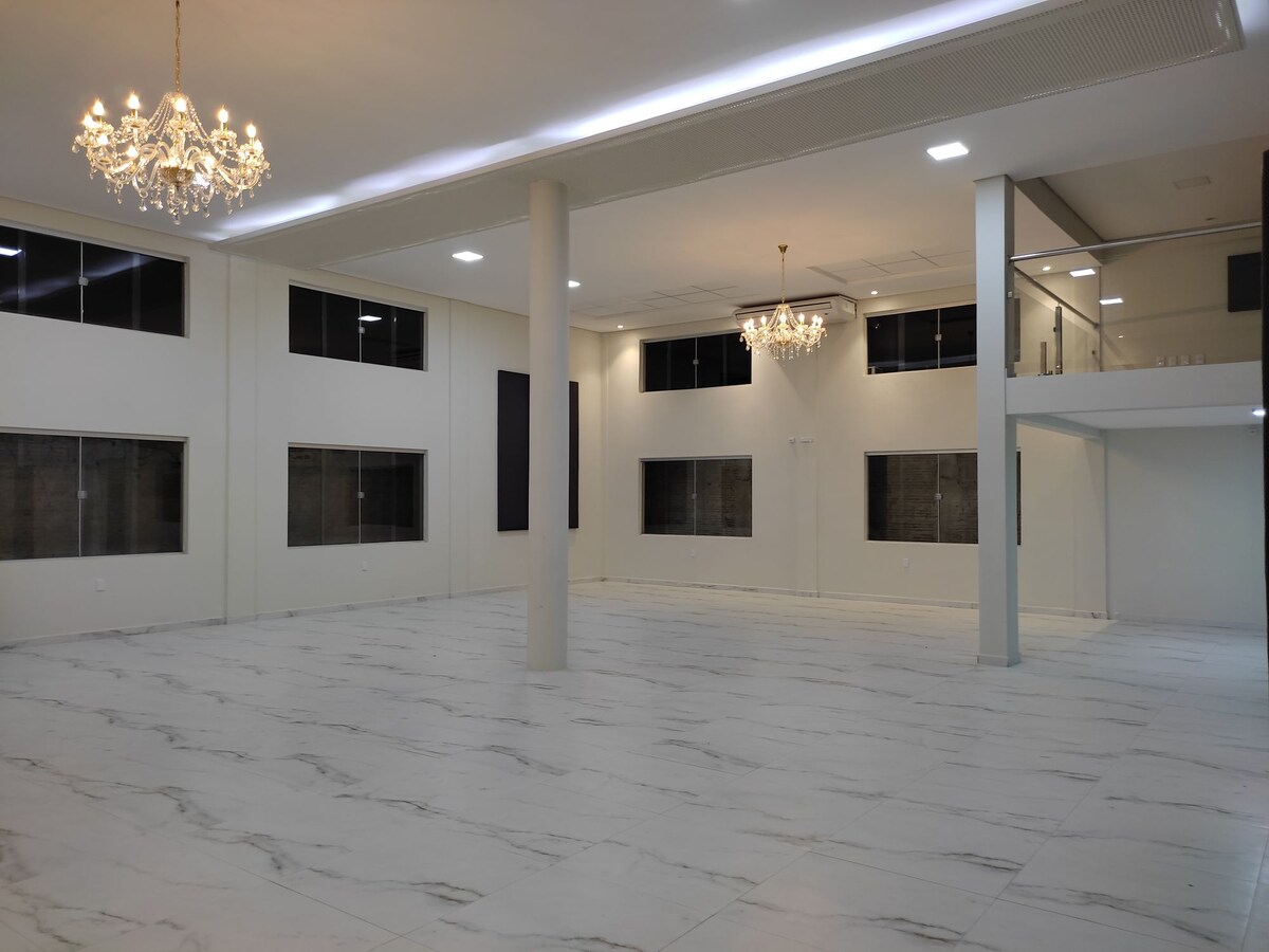 A spacious event hall features elegant chandeliers hanging from the ceiling. Large windows allow natural light to fill the area, which is accented by marble tile flooring. The layout includes an upper mezzanine that overlooks the main space, providing an open and flexible venue for gatherings.