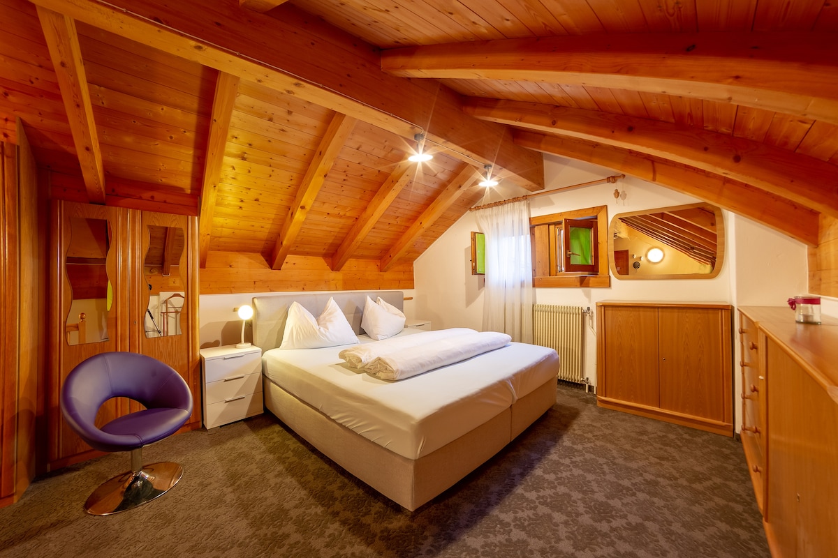 A cozy sleeping area features a spacious bed with white linens, situated beneath a wooden beamed ceiling. A modern blue chair provides seating next to a white nightstand. Natural light enters through a window with light drapes, illuminating the warm wooden accents throughout the room.