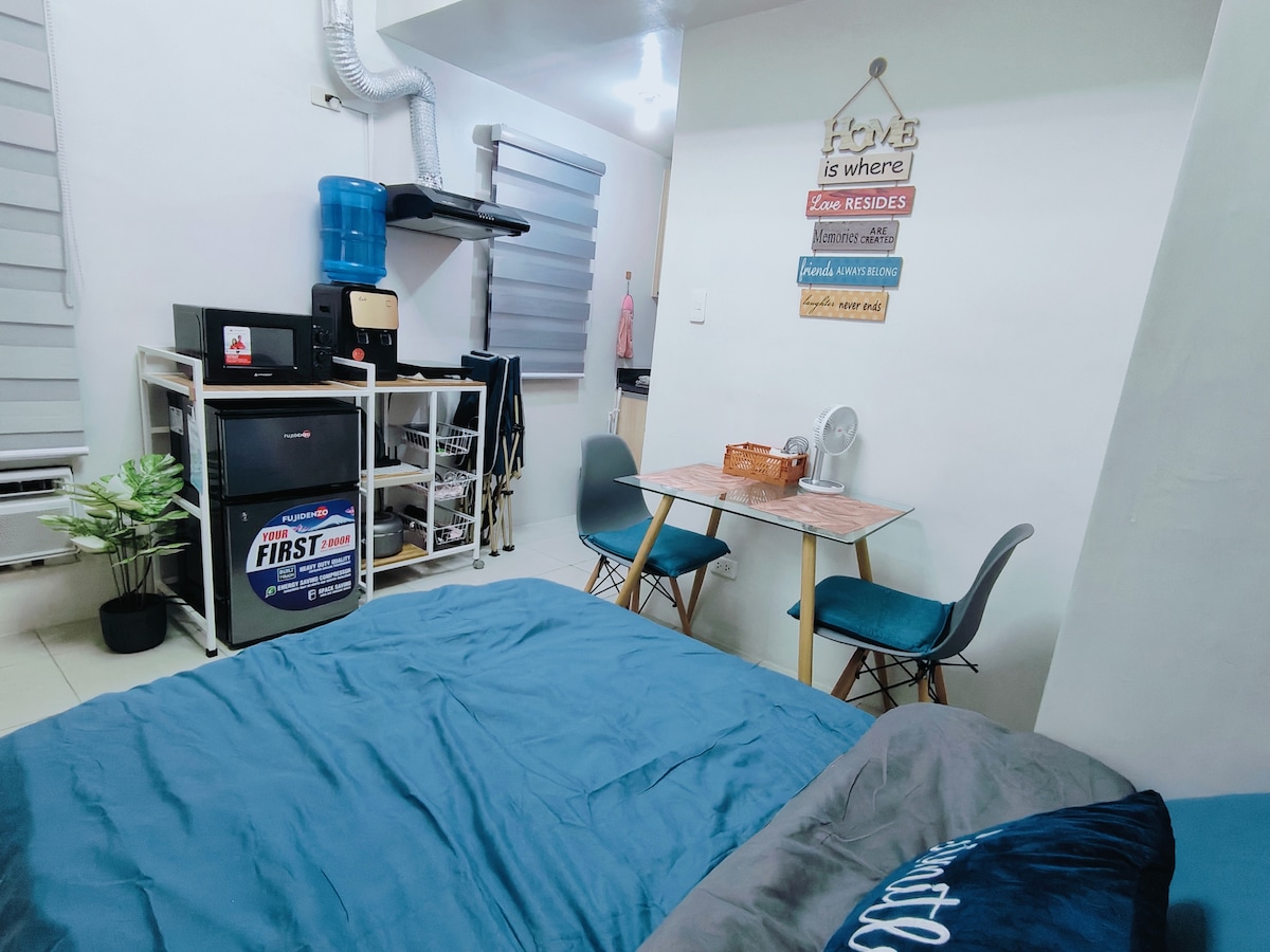 The cozy studio unit features a spacious bed with soft bedding in the foreground. A small dining table with two chairs is positioned nearby, and a kitchenette with essential appliances is visible. A decorative sign on the wall adds a personal touch to the room.