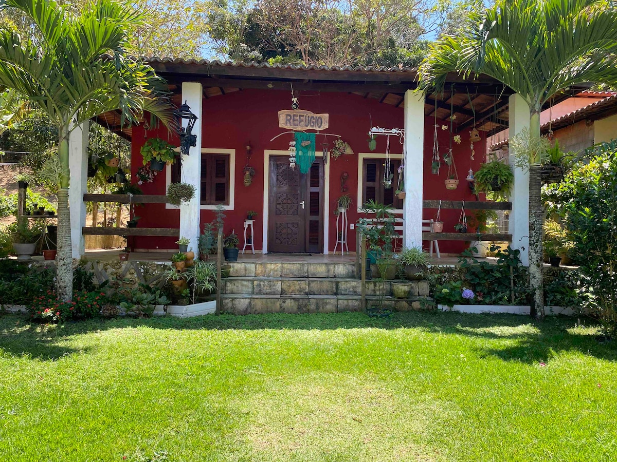The front of the house showcases a vibrant red exterior with a welcoming entrance framed by white columns. Lush greenery, including palm trees and various plants, surrounds the home, and a stone pathway leads up to the entrance, enhancing the tranquil garden setting.
