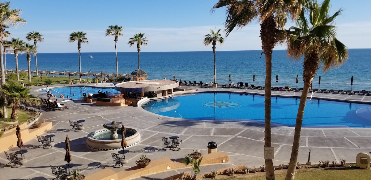 A large outdoor pool area is visible, with a swim-up bar and lounge chairs arranged alongside. Palm trees surround the space, framing the ocean view. The pool features a circular section with intricate tile designs. A few guests can be seen enjoying the area under a clear blue sky.