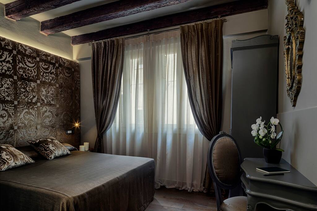 The room features a spacious bed with a soft, textured headboard. Natural light filters through a large window, framed by flowing curtains. An elegant chair and a small table complement the decor, which includes refined wall treatments and wooden beams overhead.