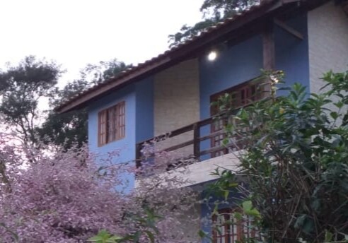A two-story building is depicted, featuring blue and light-colored walls. Balconies with wooden railings provide a view of the surrounding greenery, including blooming trees with pink flowers in the foreground. Several large trees enhance the natural setting.