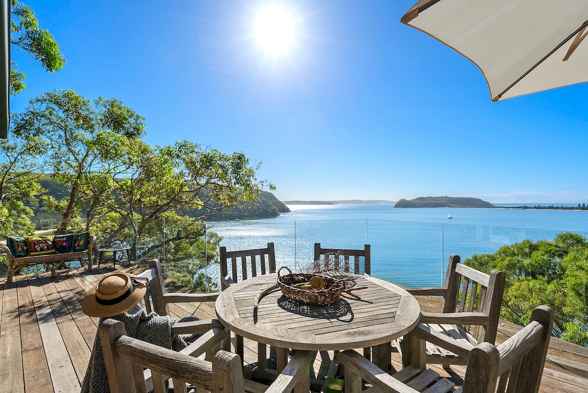 A wooden dining table surrounded by chairs is set on a sunlit deck, offering panoramic views of the water. The scene is enhanced by lush greenery and the sun shining brightly above, creating a serene outdoor dining space.