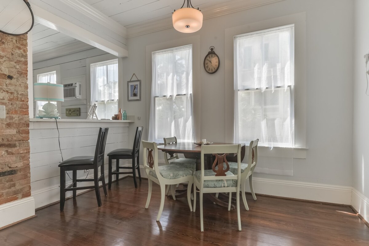 A dining area is furnished with a round table surrounded by six light-colored chairs. Natural light filters through sheer window treatments, complementing the hardwood flooring. A small clock hangs on the wall, adding to the room's inviting atmosphere.