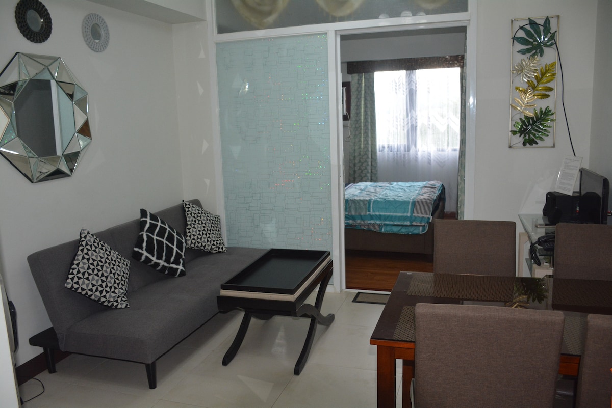 A cozy living area is observed, featuring a gray sofa adorned with patterned cushions and a sleek coffee table. A dining table with four chairs is positioned nearby. Glass sliding doors lead to a brightly lit bedroom, decorated with light curtains and a turquoise bedspread.