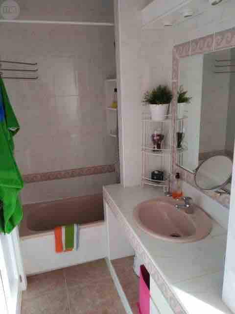 A clean bathroom is presented, featuring a bathtub with a shower above. A white sink with a mirror is positioned on a smooth countertop. Several shelves display personal care items, while towels in vibrant colors hang from a rack. Natural light enters through a nearby window.