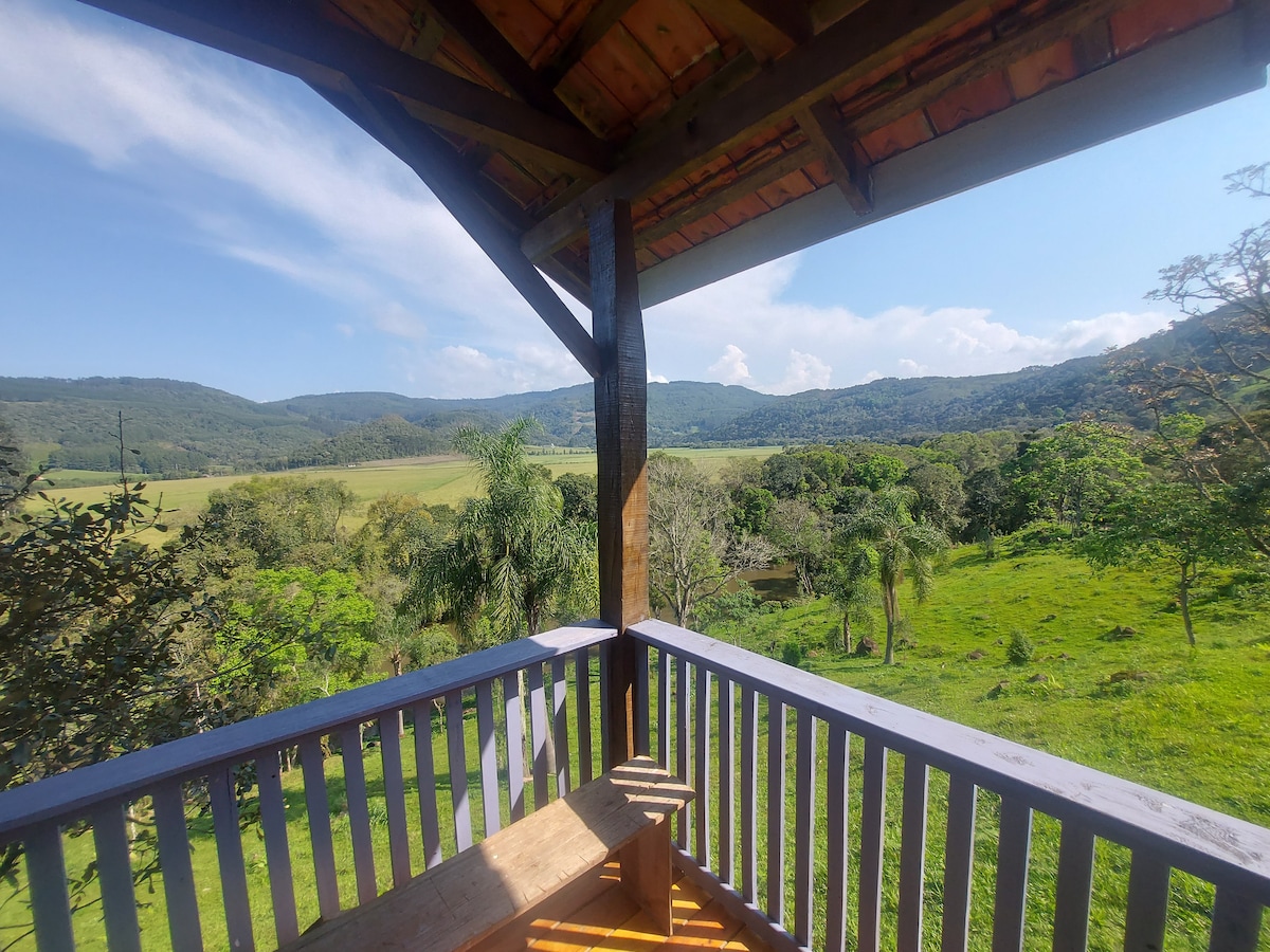 A wooden balcony offers a view of rolling green hills and distant mountains under a clear blue sky. Lush trees frame the scene, while soft grass extends into the distance, creating a serene atmosphere. The balcony railing adds a rustic touch to the expansive landscape.