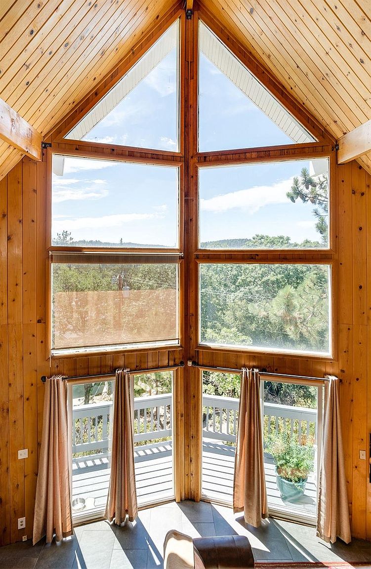 Tall, triangular windows dominate the wall, allowing abundant natural light to fill the space. Wooden paneling adds warmth to the interior, while a view of lush greenery is visible beyond the balcony. Light, flowing curtains frame the windows softly.