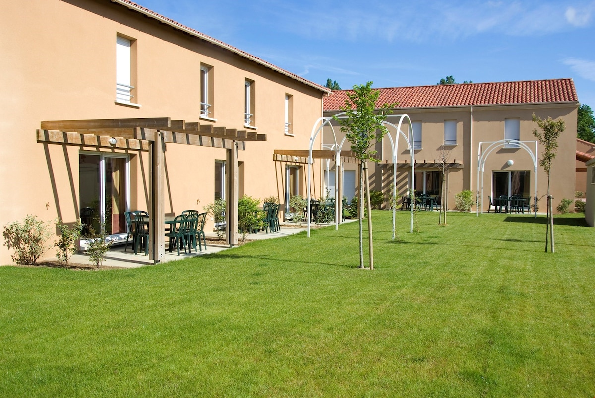 A spacious garden area features well-maintained grass and several newly planted trees. Multiple patio areas with dark green chairs and tables are visible, set against the light stone facade of the buildings, enhancing the inviting outdoor space.