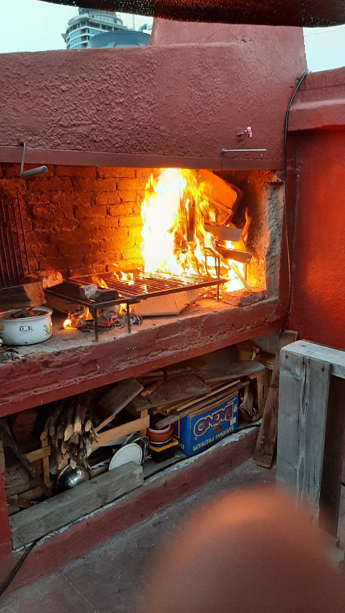 An outdoor barbecue area features a brick grill with flames and glowing embers. The space is surrounded by exposed wood storage, outdoor utensils, and a cozy rustic design. Warm light reflects off the surfaces, creating a welcoming environment for gatherings.