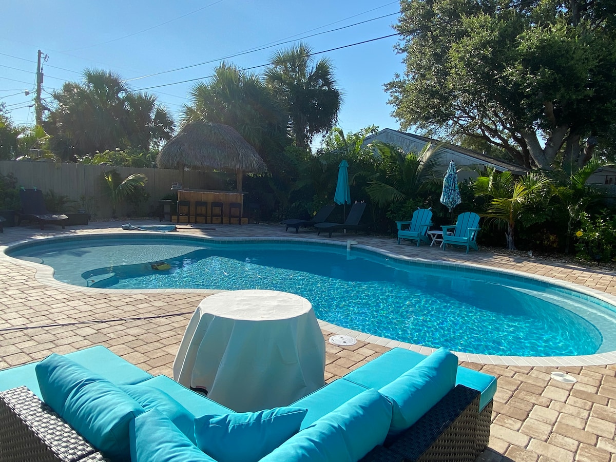A tropical backyard features a sparkling heated pool surrounded by lush greenery. Lounge chairs in vibrant blue offer relaxation, while a tiki bar provides additional seating. Sun umbrellas add shade, and a small table is positioned near the pool, creating an inviting outdoor space.