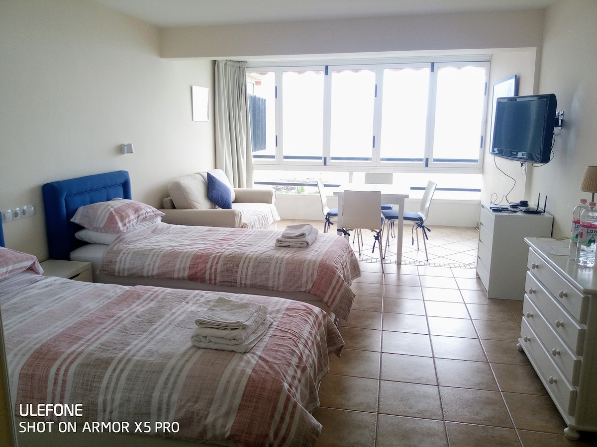 A comfortable studio space features two twin beds with soft linens and a nightstand beside each. A cozy seating area is included, along with a dining table and chairs. Large windows offer abundant natural light, showcasing views of the surrounding area.