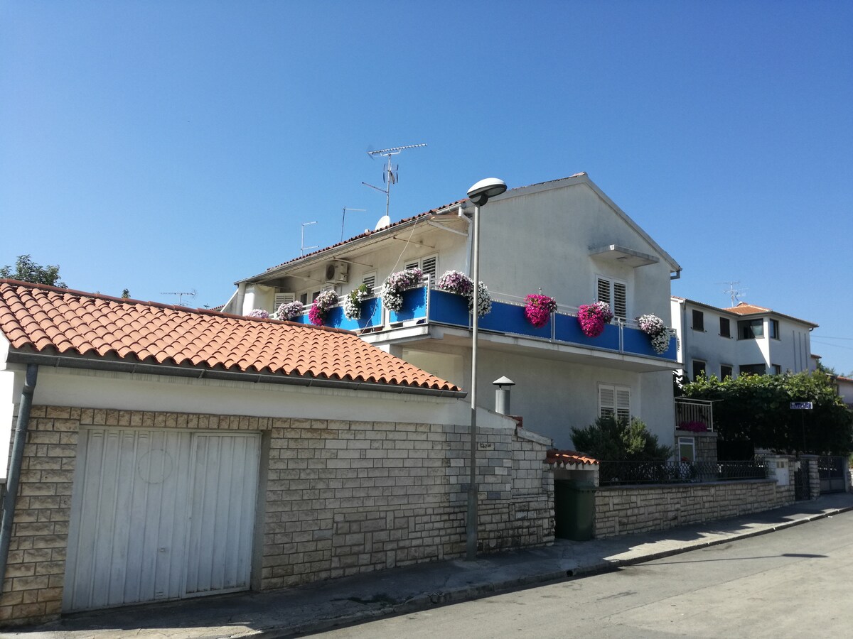 A multi-level building is shown, featuring a white exterior with a red-tiled roof. Flower boxes adorned with colorful blooms are visible on the upper balcony. A single-story structure next to it includes a garage with a wooden door, and street elements like a lamp post are present.