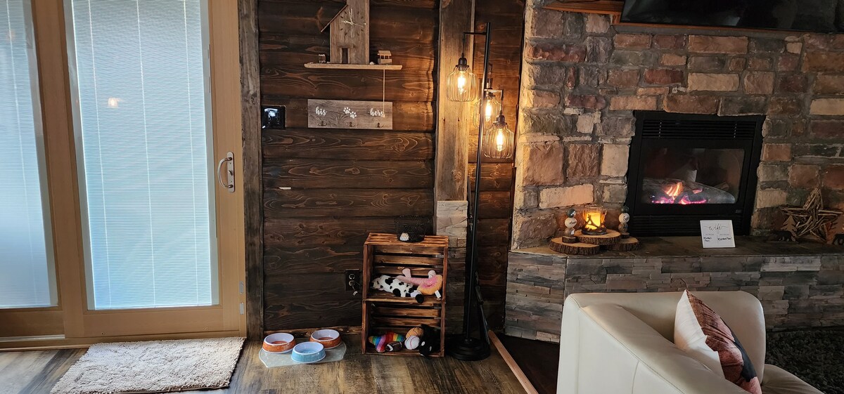 A cozy living area features a stone fireplace, surrounded by wooden accents. A decorative shelf holds various items, while a selection of pet bowls is neatly arranged below. Soft lighting from the stylish floor lamp adds warmth to the space, complemented by the natural light from the glass doors.