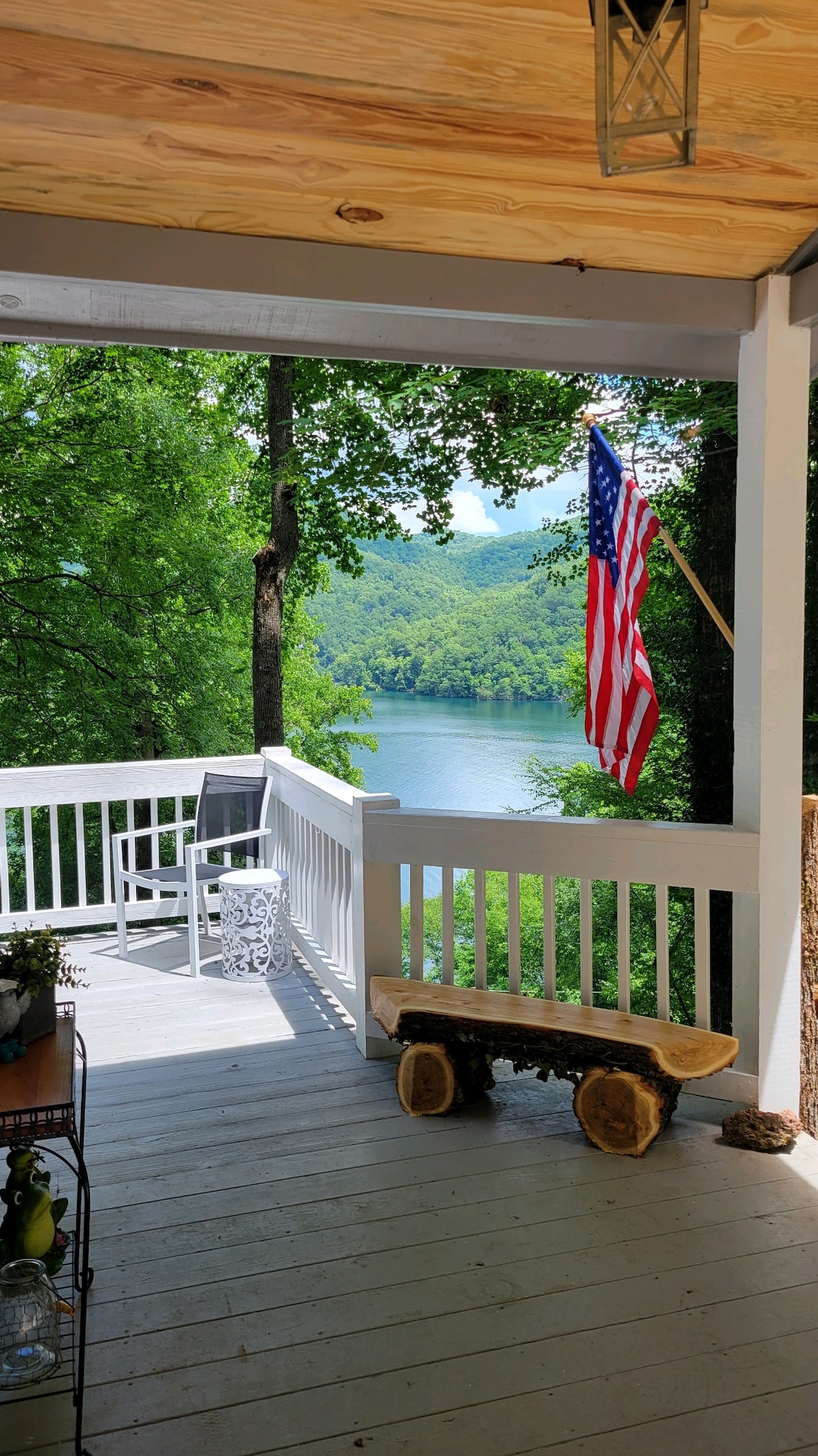 A spacious outdoor deck is featured, overlooking a serene lake surrounded by lush greenery. An American flag gently waves in the breeze, while a wooden bench and a white chair offer cozy spots to enjoy the view. Sunlight filters through the leaves, enhancing the tranquil ambiance.