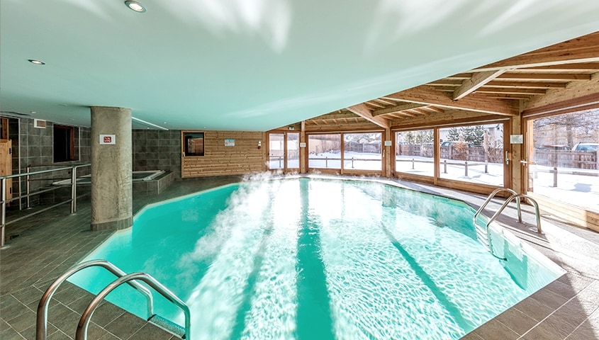 An indoor pool is shown, featuring clear turquoise water with gentle steam rising. Surrounding windows allow natural light to fill the space, while wooden beams enhance the architectural design. Accessible steps lead into the pool area, emphasizing a relaxing environment.