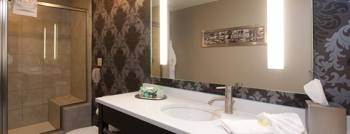 A modern bathroom features a spacious walk-in shower with glass doors and textured tile walls. An elegant vanity with a large mirror is complemented by contemporary lighting. A decorative wall showcases floral wallpaper, while plush towels and complimentary toiletries are neatly arranged on the counter.