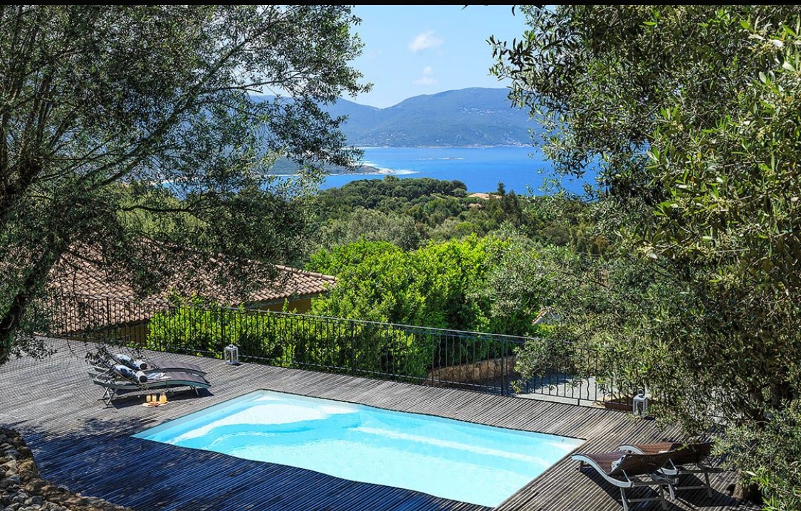 A refreshing heated pool is framed by wooden decking and surrounded by lush greenery. It offers a clear view of the blue waters of the Gulf of Valinco and the landscape beyond, inviting tranquility and relaxation.