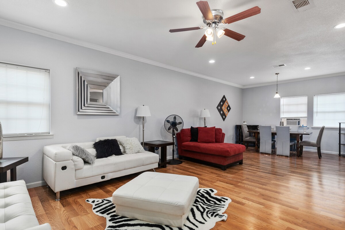A spacious living area features hardwood flooring and a ceiling fan. A white sectional sofa and a red couch are situated near a coffee table. A dining area with a table and chairs is visible in the background, accompanied by two windows allowing natural light.