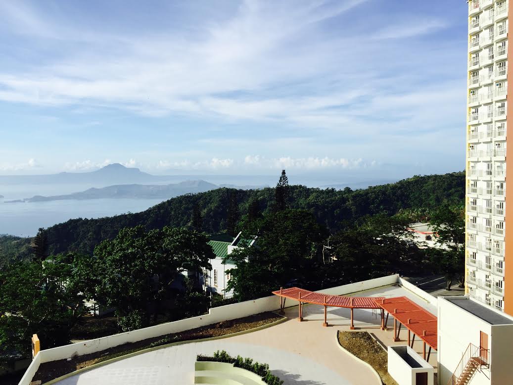 A panoramic view of Taal Lake and surrounding mountains is captured from the balcony. Lush greenery adorns the landscape, while the clear sky and distant volcanic peaks provide a serene backdrop. The balcony area includes shaded seating under a trellised structure.