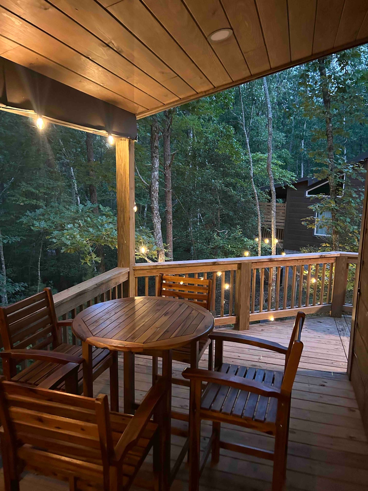 A wooden deck is featured with a round table and four matching chairs, illuminated by string lights. The surrounding trees offer a natural backdrop, enhancing the secluded and tranquil atmosphere.