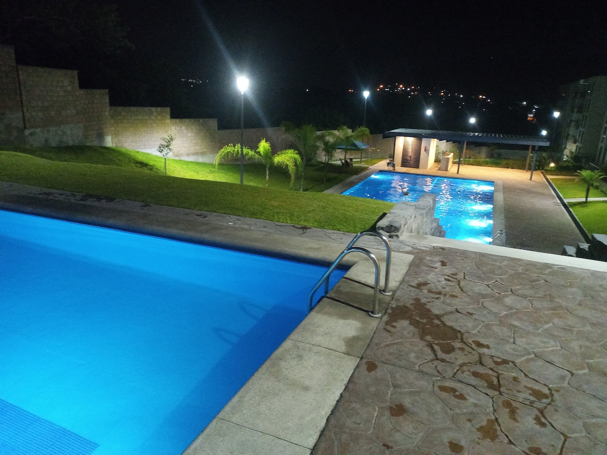 Two illuminated pools are visible under the night sky, surrounded by lush grass and neatly arranged palm trees. The pools offer a calming blue hue, while the area is softly lit by nearby lights and offers scenic views in the background.