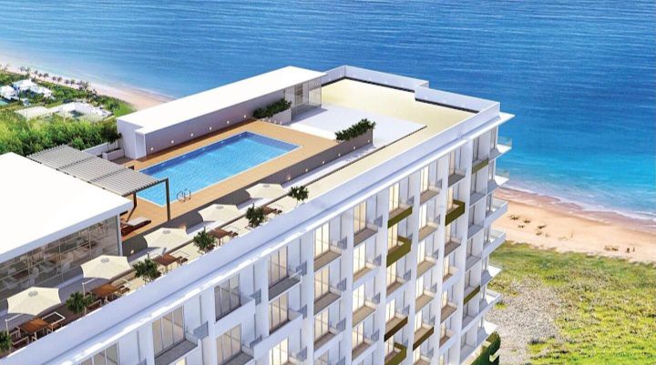 An aerial view captures a modern beachfront building, showcasing a rooftop pool surrounded by lounge areas. The distant shoreline and ocean are visible, with a serene beach enhancing the environment. Bright sunlit spaces and neatly arranged outdoor furniture are noticeable on the terrace.