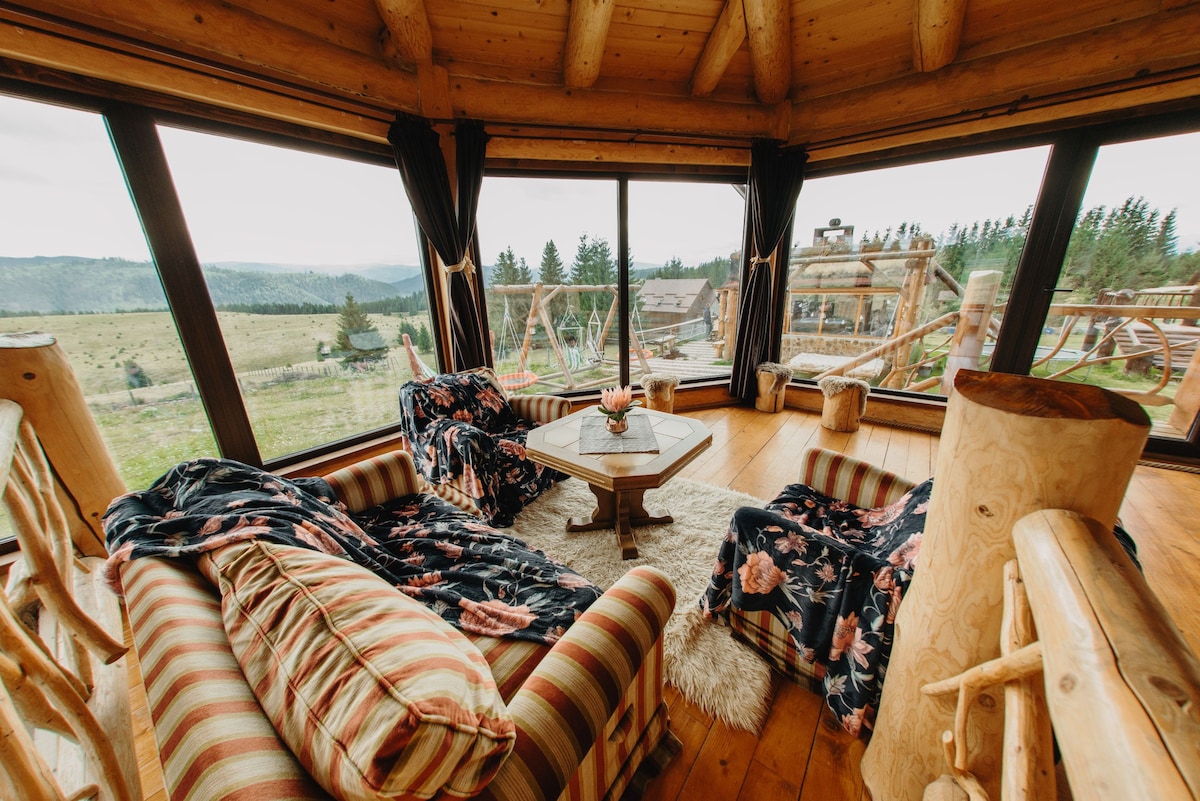 A cozy seating area features two plush lounge chairs adorned with floral covers, positioned around a wooden coffee table. Large windows provide expansive views of the surrounding landscape, while natural wood accents and a soft area rug contribute to the room’s welcoming environment.