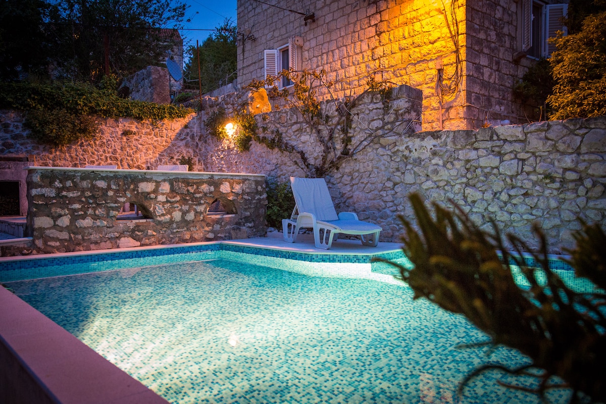 A tranquil outdoor space features a stone wall illuminated by a warm light. A swimming pool with a textured surface reflects surrounding greenery. Two lounge chairs are positioned beside the pool, providing a serene area for relaxation.