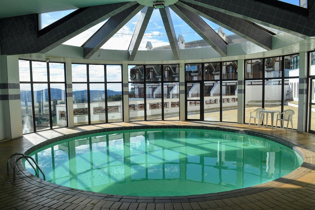 An enclosed swimming pool area features a circular pool with clear water under a skylight roof. Large glass panels create a bright space, allowing views of the surrounding landscape. Simple white chairs are placed near the pool, enhancing the open and airy atmosphere.