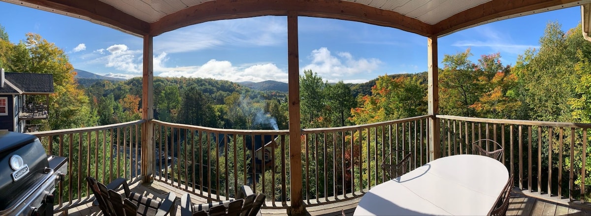 A panoramic view from a private balcony showcases trees transitioning through autumn colors, framed by a clear blue sky. A table and chairs are positioned for relaxation, while a grill is visible, ideal for outdoor dining. The serene landscape invites tranquility.