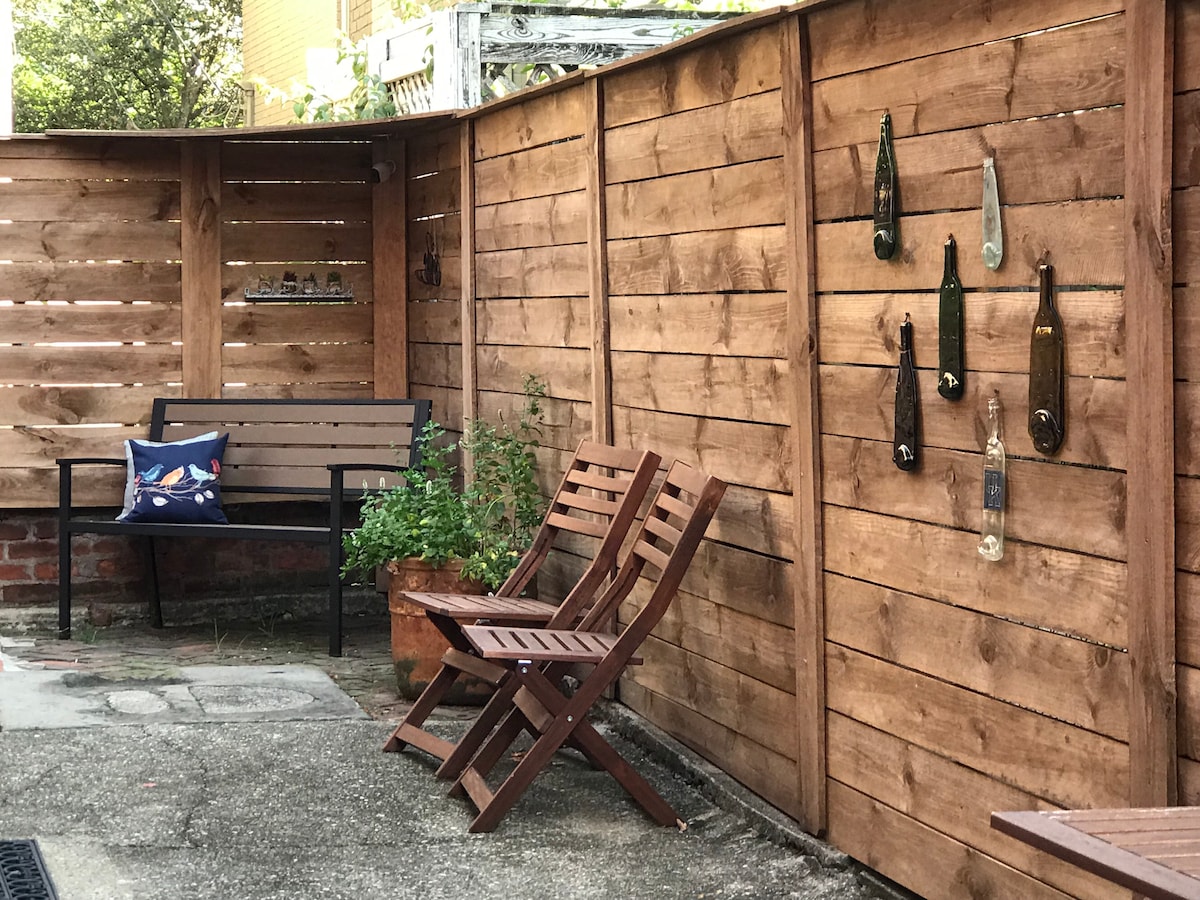 An outdoor patio area features wooden fencing and two seating options: a black metal bench and a pair of wooden chairs. Decorative glass bottles are mounted on the fence, with a potted plant nearby, adding a touch of greenery to the space.
