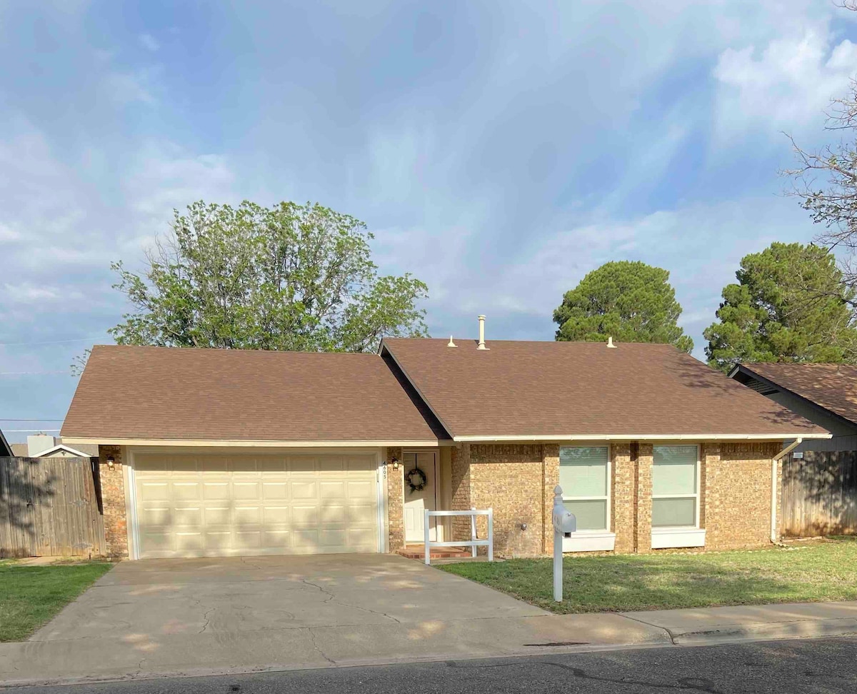 A single-story home with a brown shingle roof and brick exterior is set against an overcast sky. A double car garage is visible on the left, while well-maintained grass surrounds the driveway. Mature trees provide a natural backdrop in the residential neighborhood.