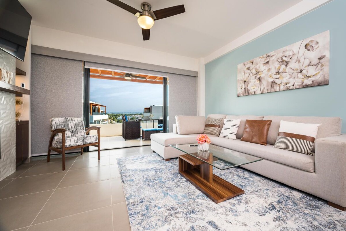 A stylish living area features a large sectional sofa in soft tones, complemented by decorative pillows. A glass coffee table sits atop a textured area rug. Expansive sliding doors open to a balcony, inviting natural light and a view of outdoor space.
