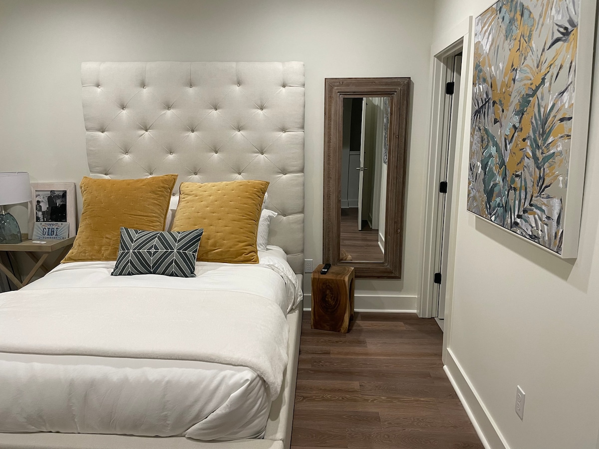 A bright bedroom features a light-colored upholstered headboard, adorned with two decorative yellow pillows and a geometric black and white cushion on a spacious bed. A wooden mirror and a piece of wall art with botanical motifs can be seen to the side.