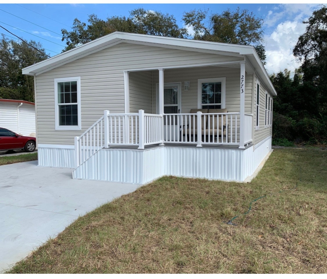 A light-colored cottage with a spacious porch, featuring white railings that lead up to the entrance. The exterior is complemented by manicured grass and clear blue skies, while parking spaces for vehicles are visible nearby.