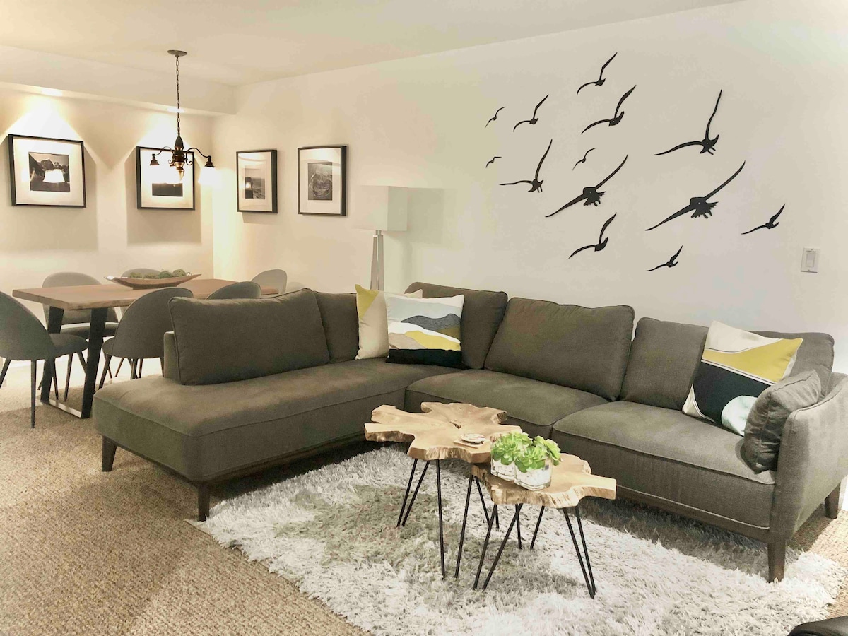 A spacious living area features a gray sectional sofa adorned with decorative cushions. A natural wood coffee table sits on a textured rug. Framed photographs are displayed on the wall, and a dining table with modern chairs is visible in the background.