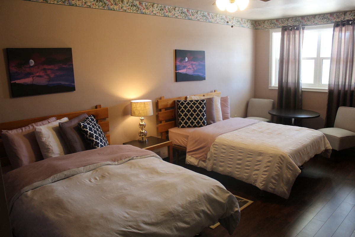 A spacious room contains two beds with neutral-colored linens. A small round table is accompanied by two chairs. Soft lighting from a lamp complements natural light filtered through sheer curtains on large windows. Floral wallpaper adds a subtle decorative touch.