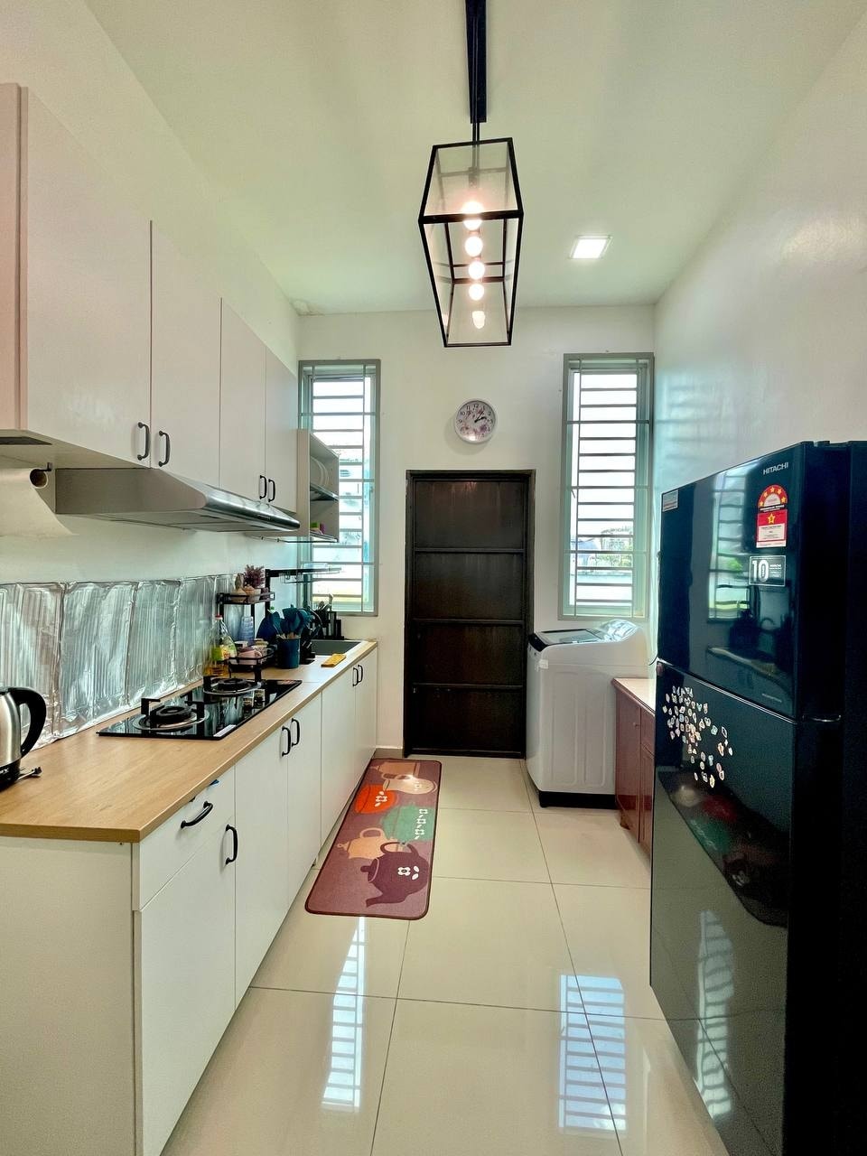 A modern kitchen is equipped with essential appliances, including a fridge and stove. Natural light enters through two windows, complementing the clean design. A decorative mat adds a touch of color to the tiled floor, while overhead lighting enhances visibility.