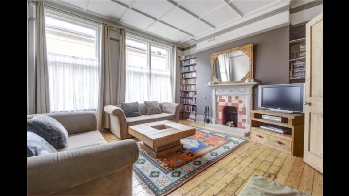 A spacious living room is characterized by high ceilings and natural light from large windows. A central coffee table rests on a decorative rug, surrounded by a comfortable sofa and chairs. A traditional fireplace serves as a focal point, accompanied by a bookshelf and television.
