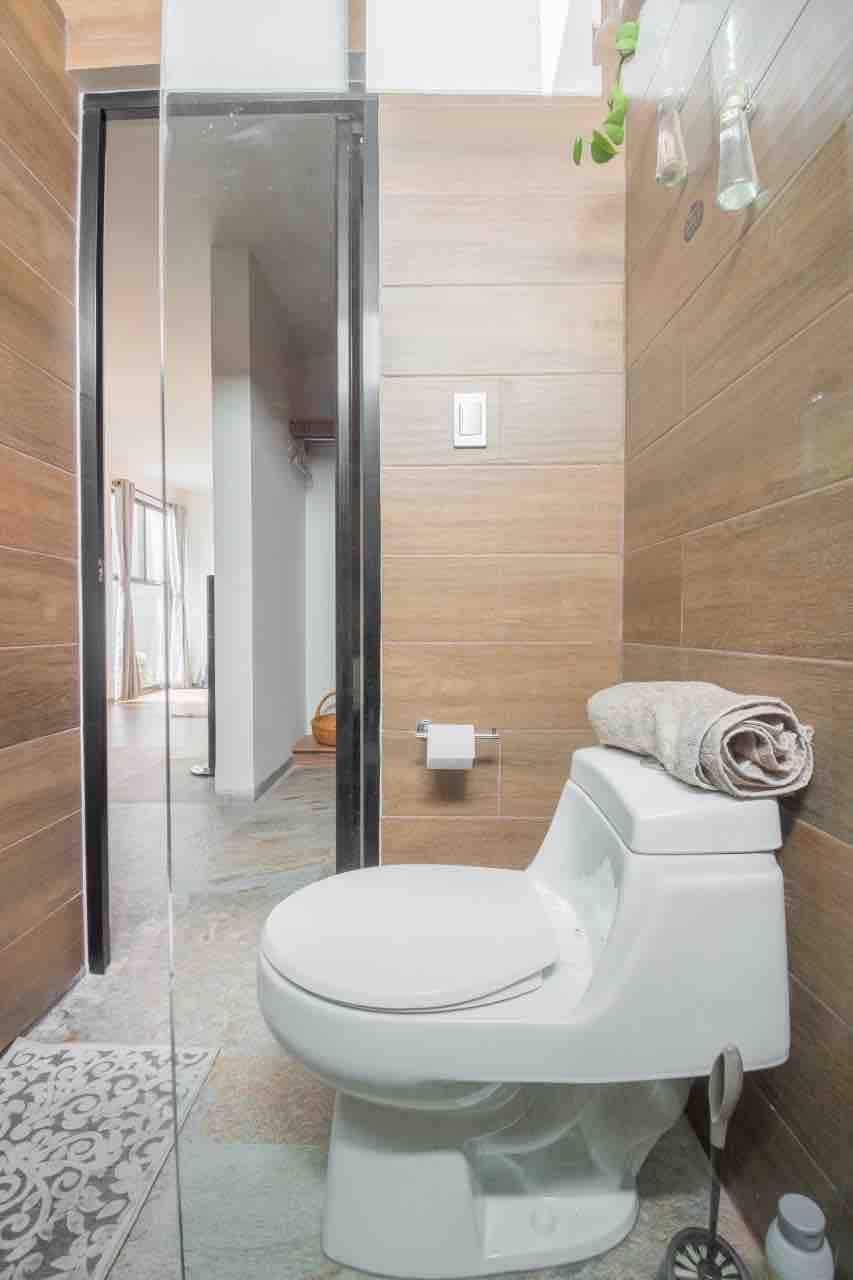 The bathroom features a modern design with tiled walls and a glass shower enclosure. A clean white toilet is positioned next to a folded towel on a rack. Natural light is visible through the door leading to the remaining living space.