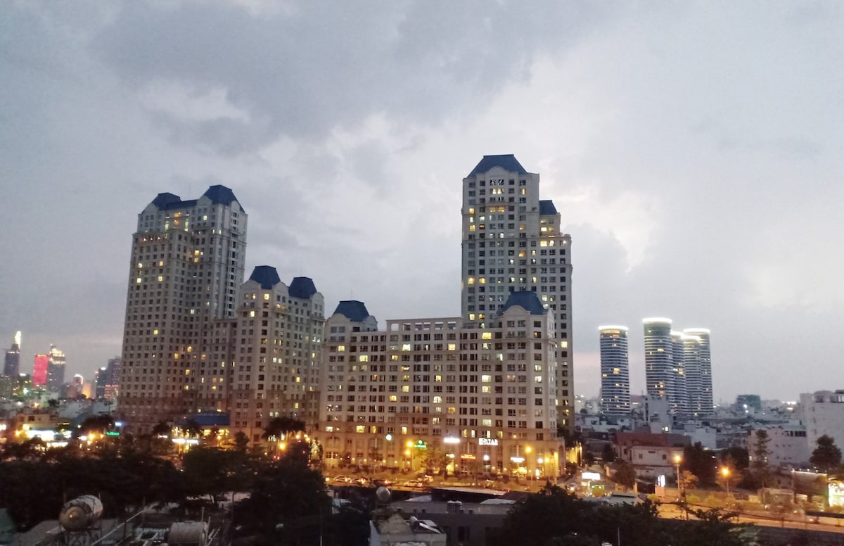 The image captures a view of modern high-rise buildings under a cloudy sky. The structures feature distinctive blue rooftops and numerous windows reflecting city lights. In the background, city skyscrapers and hints of vibrant city life can be seen, adding to the urban landscape.