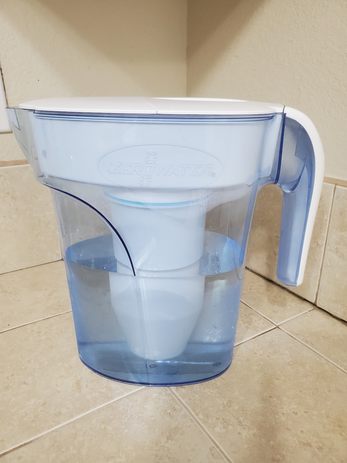 A water filtration pitcher is placed on a tiled surface, featuring a transparent blue design with a white lid and handle. Water is visible inside the pitcher, highlighting its filtration system.