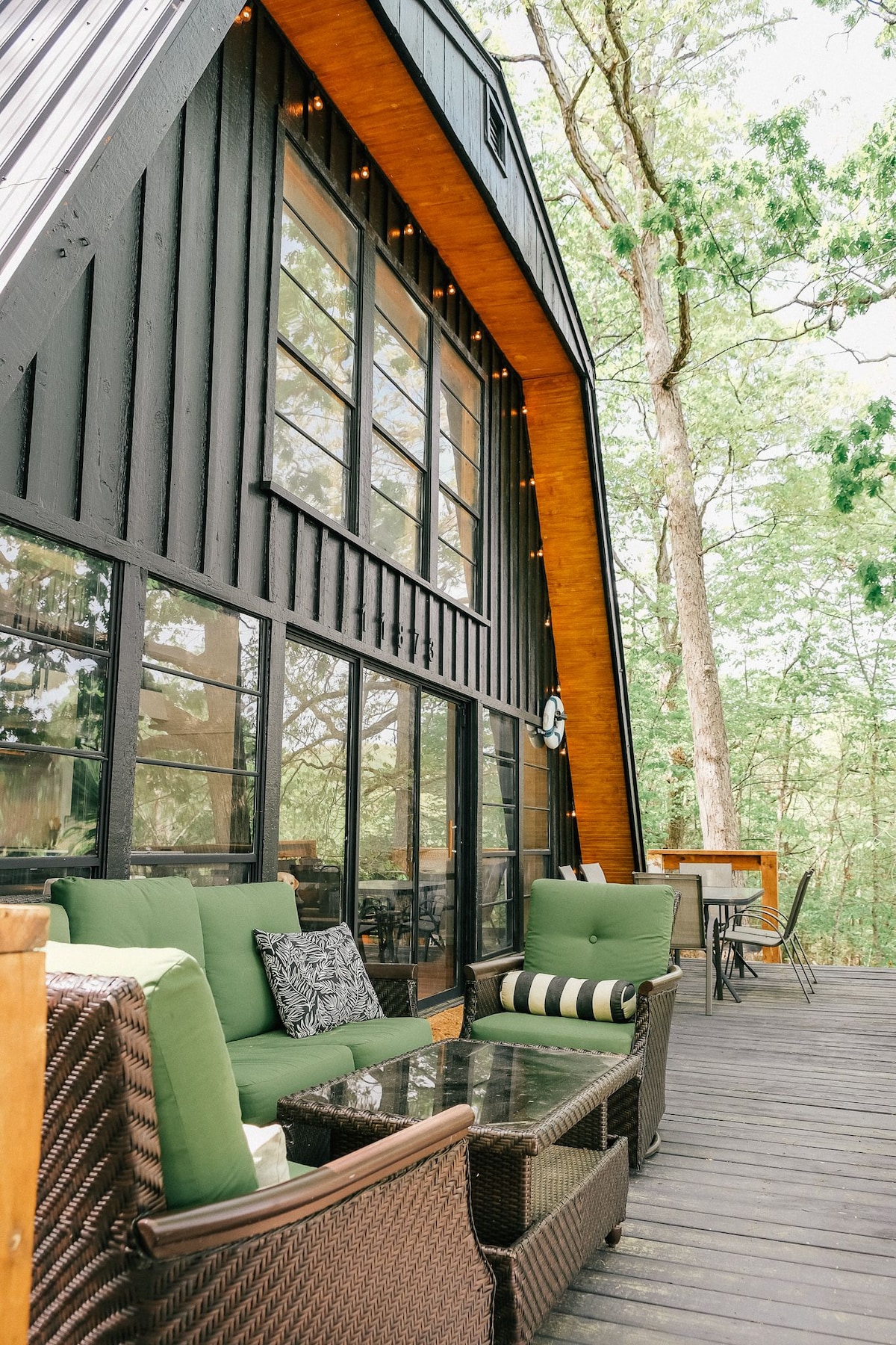 A spacious outdoor deck features comfortable seating, including green cushioned chairs and a glass coffee table. Large windows are visible, providing a connection to nature and inviting natural light indoors. Surrounding trees enhance the serene outdoor environment.