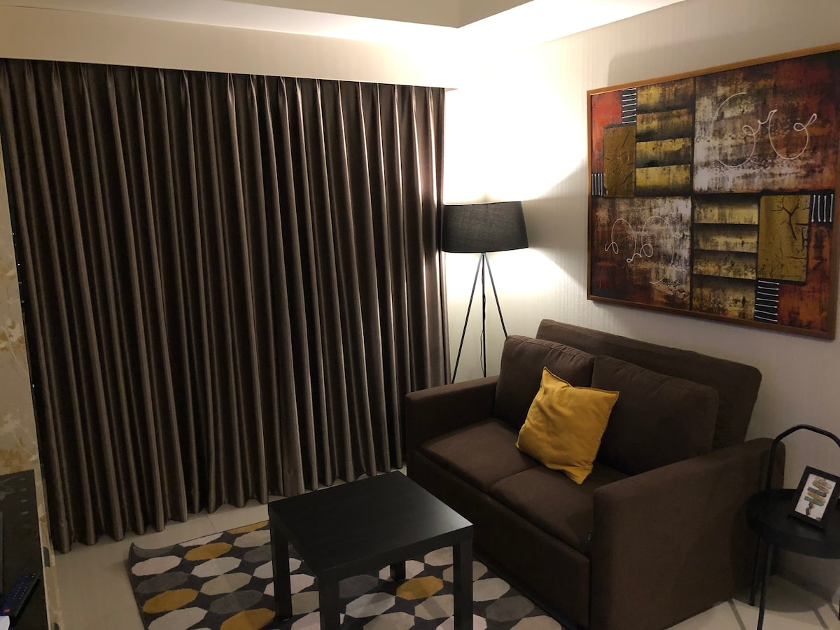 A cozy living area is featured with a dark brown sofa adorned with a yellow accent pillow. A low black table sits in front, while a patterned rug is placed beneath it. Sheer curtains frame a window, and a standing lamp provides soft lighting beside the seating.